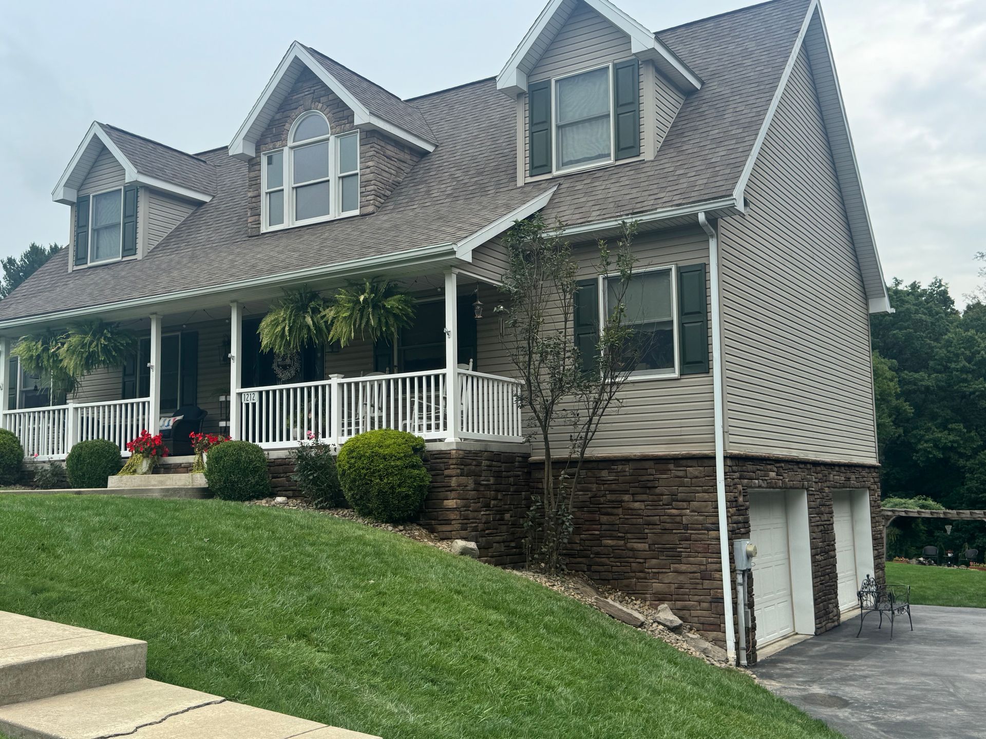 Gray two-story house with dormer windows, white porch railing, and driveway beside a sloped lawn