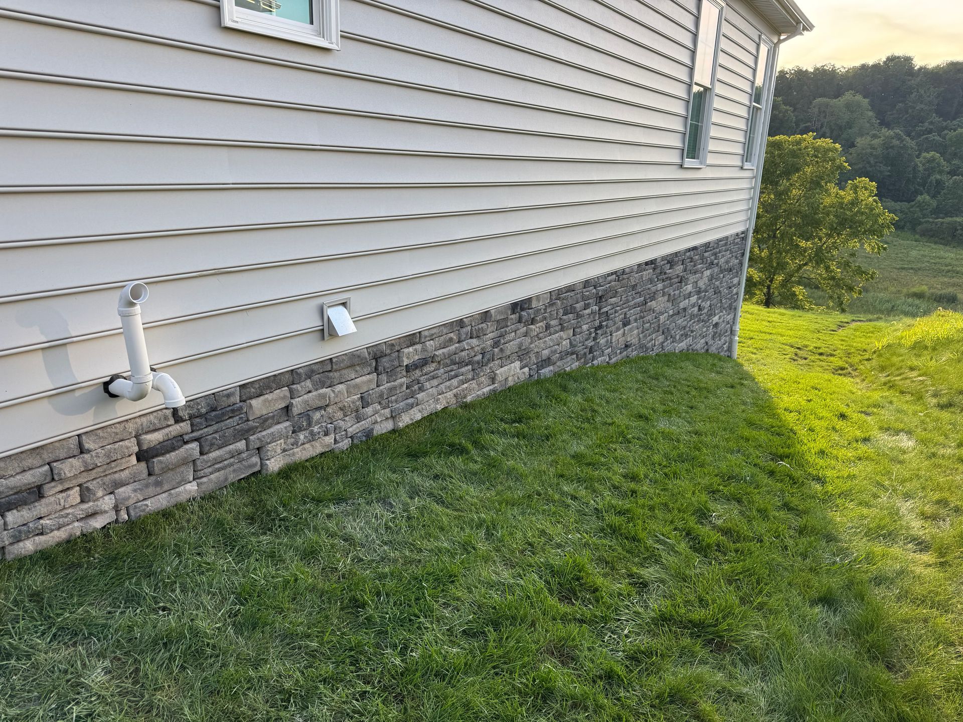 House siding and stone foundation beside a grassy yard on a sunny hillside