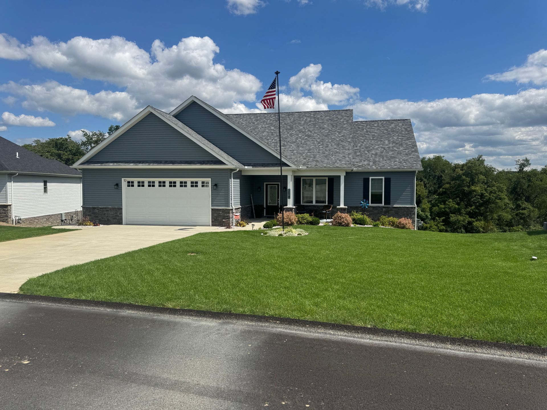 Suburban blue-gray house with garage, large lawn, and American flag under a partly cloudy sky