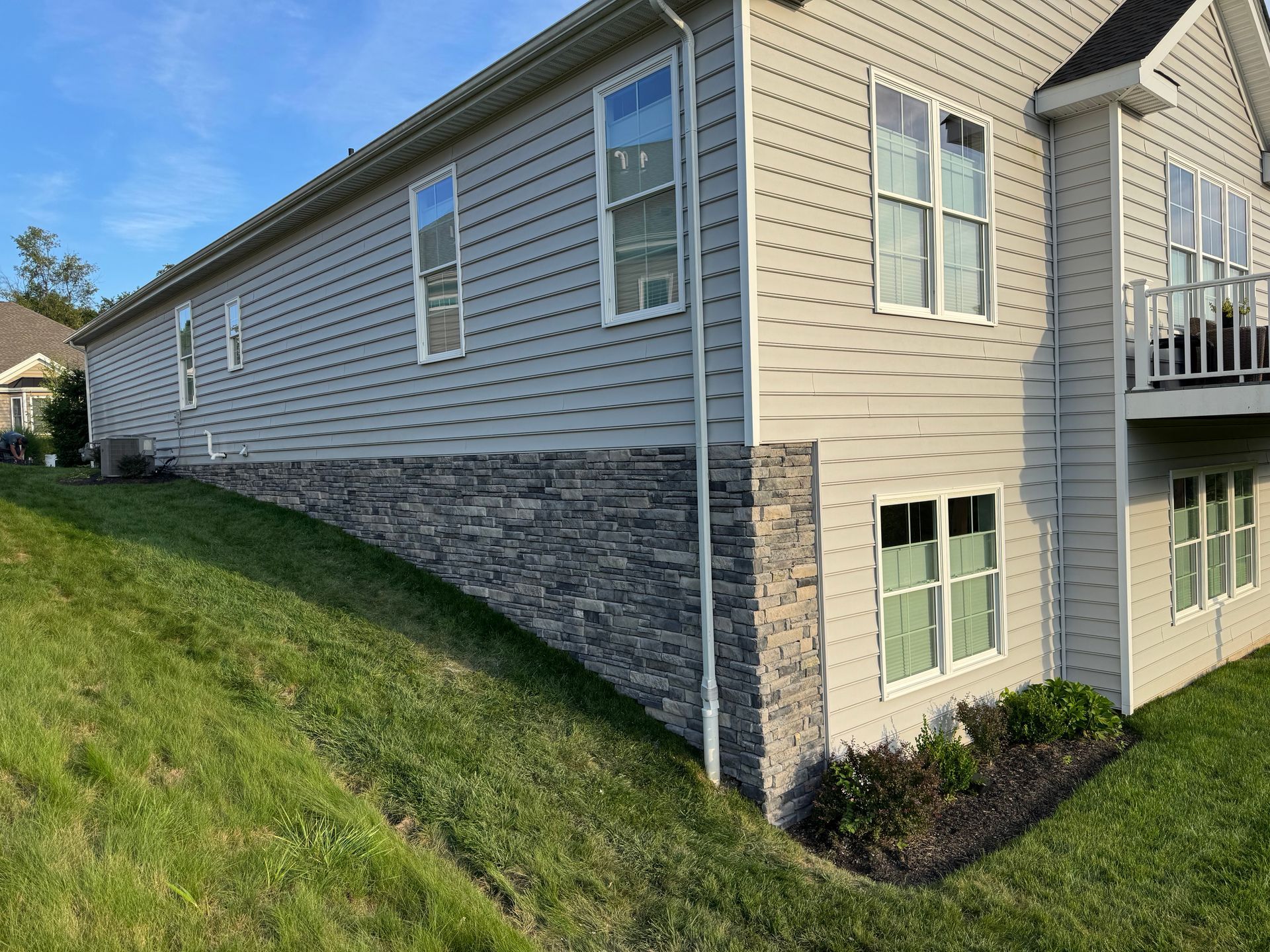 Side view of a beige house with stone foundation on a grassy slope and small windows.