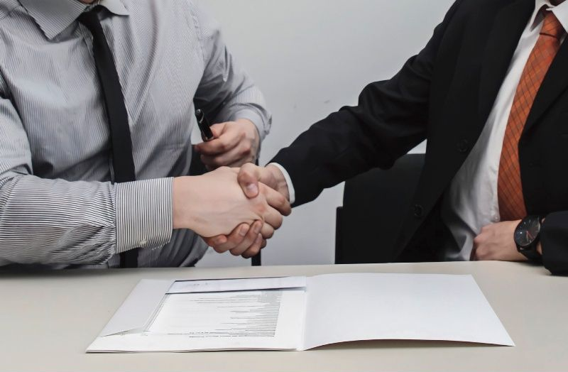 Two men in suits shaking hands over a document on a table.