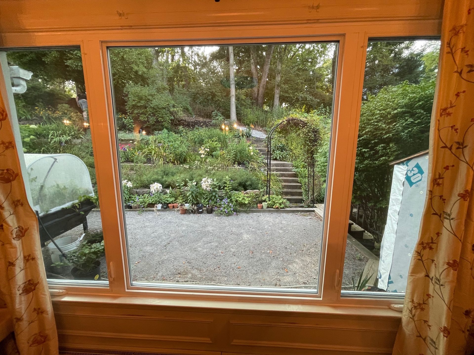 View through a wooden window to a gravel patio and lush garden with stone steps and greenery