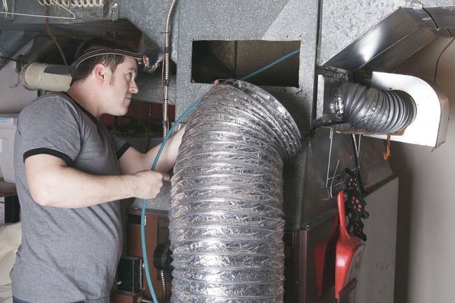 Technician inspecting HVAC ductwork in a basement, holding a tool beside shiny flexible ducts.