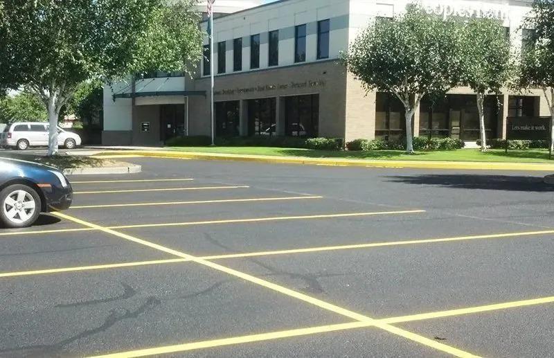 Parking lot in front of a tan building with trees and parked cars. Yellow lines mark spaces.