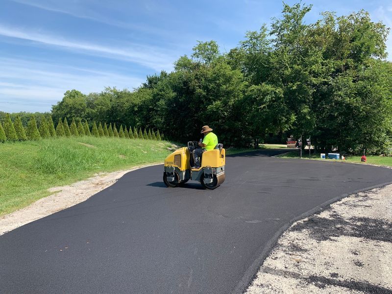 A worker on a yellow roller compacts fresh asphalt paving on a driveway under a blue sky.