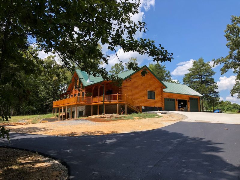Log cabin with a green roof, wraparound deck, and attached garage on a sunny day.