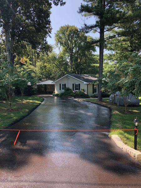 Asphalt driveway leads to a light-colored house amid trees under a blue sky.