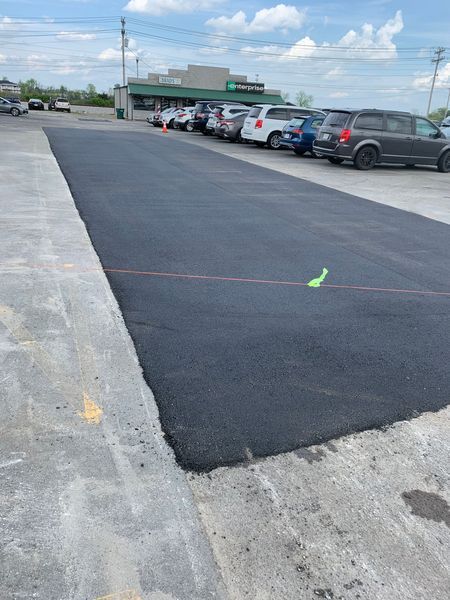 Freshly paved asphalt in a parking lot, with parked cars and a building in the background.