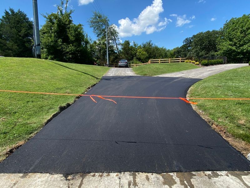 Newly paved asphalt driveway curving between green lawns; vehicle in the background.