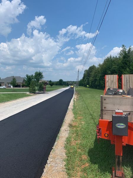 Newly paved road next to grassy area, with a utility trailer on the right under a blue sky.