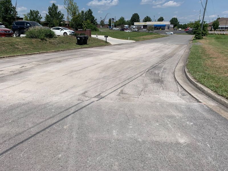 Paved road with light-colored markings, curved sidewalk on right, cars, trees and buildings in the background.