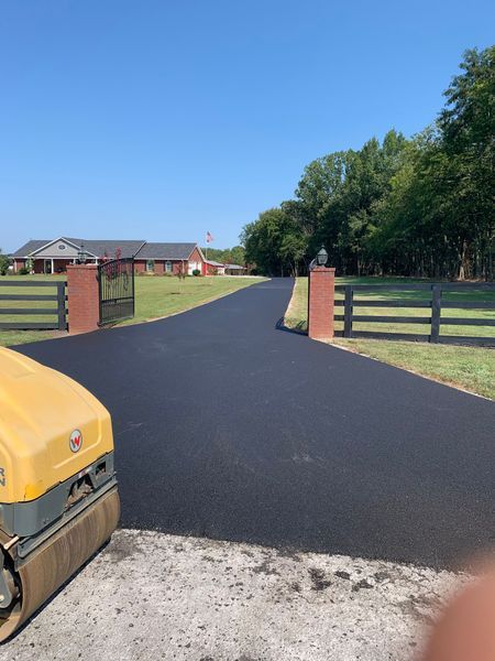 Asphalt driveway, freshly paved, leads to a house. Roller visible on left. Brick gate posts.