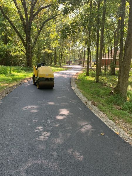 Asphalt paving machine on a newly paved road in a wooded area.