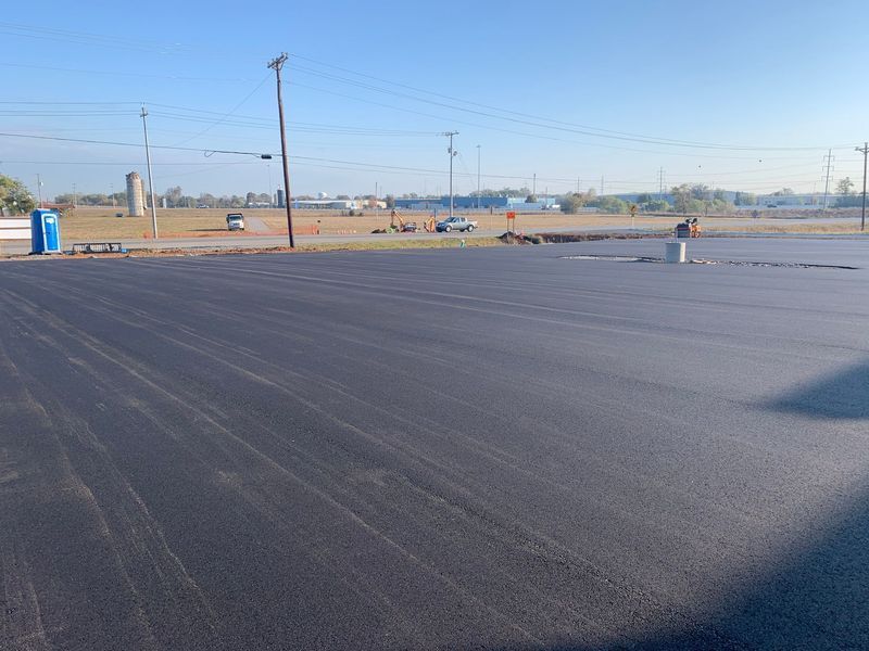 Newly paved asphalt lot with construction in the background under a clear, blue sky.