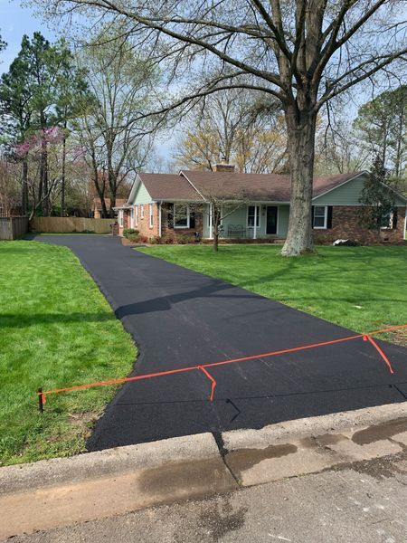 Black asphalt driveway in front of a brick and siding house with green lawn and trees.