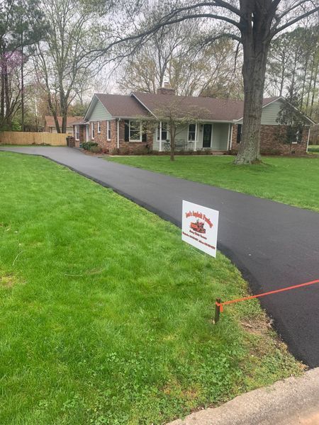 Newly paved asphalt driveway leading to a brick house with a sign on the lawn.