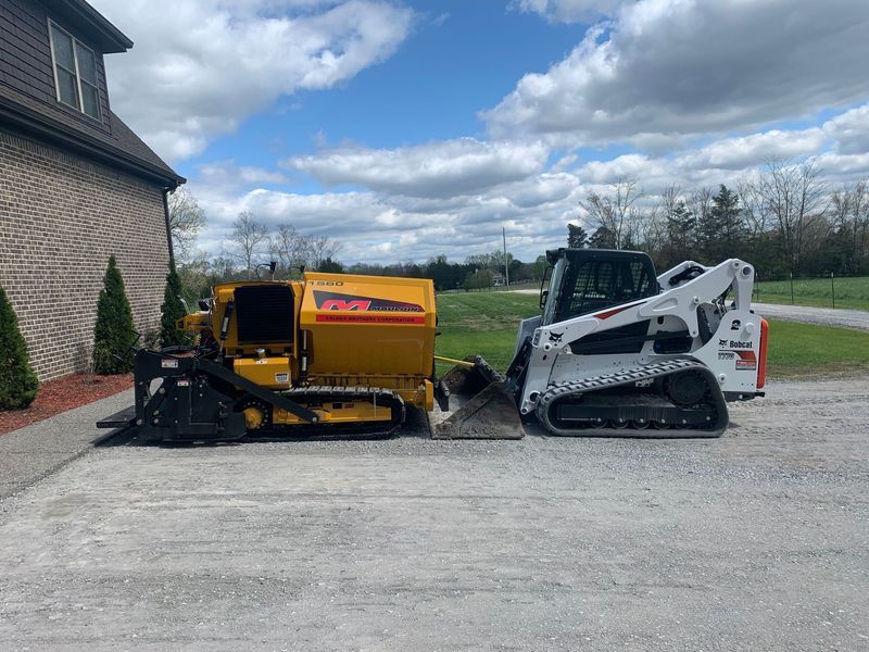 Yellow asphalt milling machine and white Bobcat skid steer on pavement beside a building under a cloudy sky.