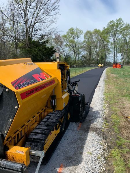 Yellow asphalt paver laying fresh black asphalt on a gravel path, other machinery in the background.