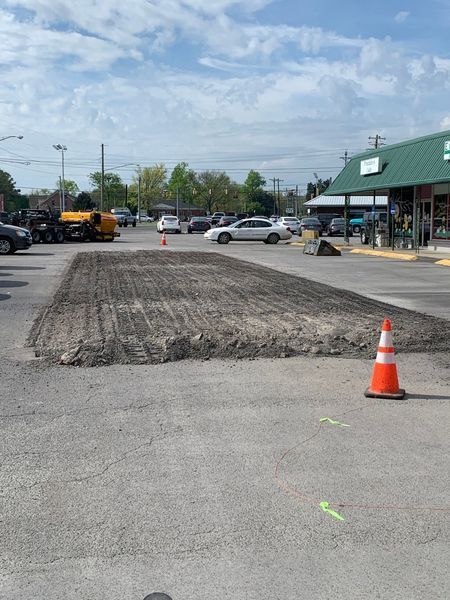 Parking lot with section of asphalt removed, cone in foreground, building and vehicles visible.