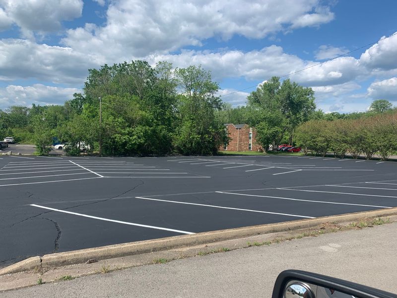 Empty asphalt parking lot with white lines; trees in the background, cloudy blue sky.