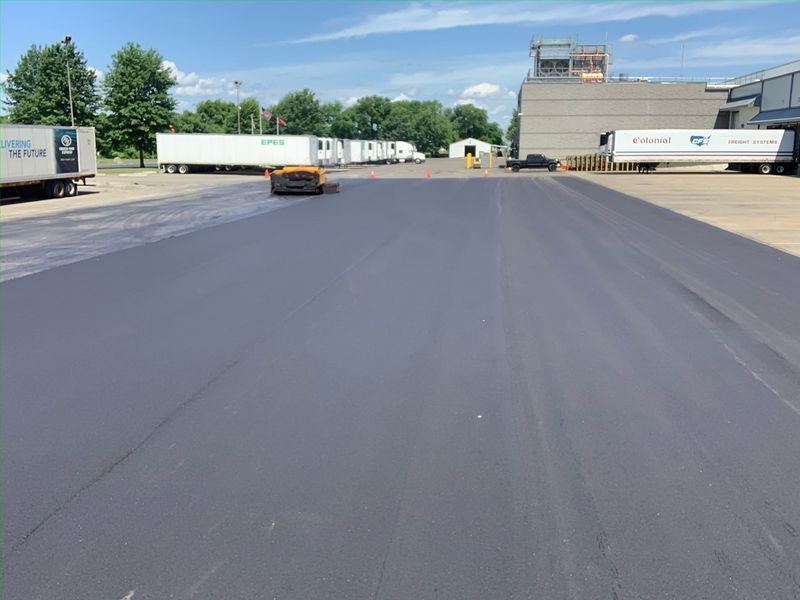 Newly paved black asphalt parking lot with parked semi-trucks and industrial buildings under a blue sky.