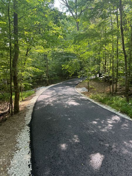 Paved path winds through a lush green forest, bordered by trees and gravel.
