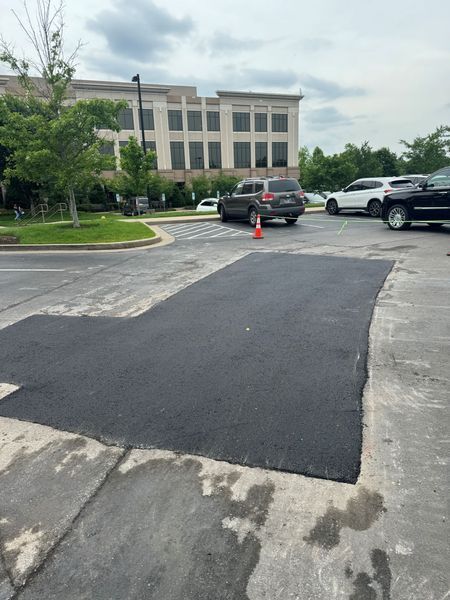 Black asphalt patch in a parking lot with cars and a building in the background.