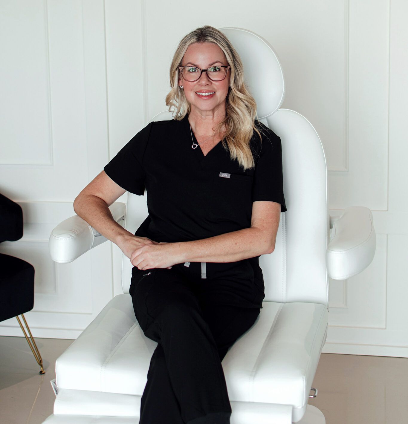 A woman wearing glasses is sitting in a white chair.