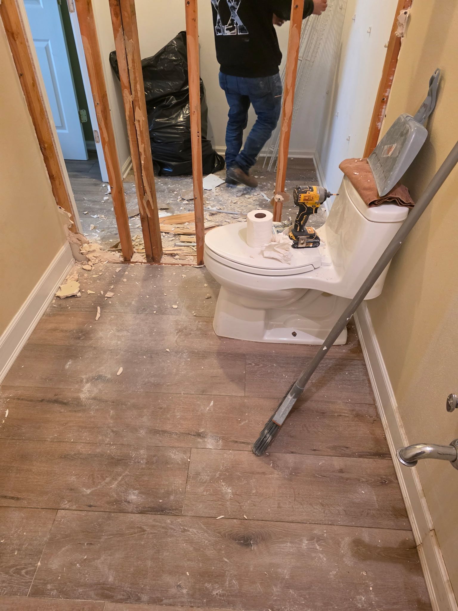 Bathroom renovation in progress toilet, drill, dust, and debris are visible. A person works in the background.