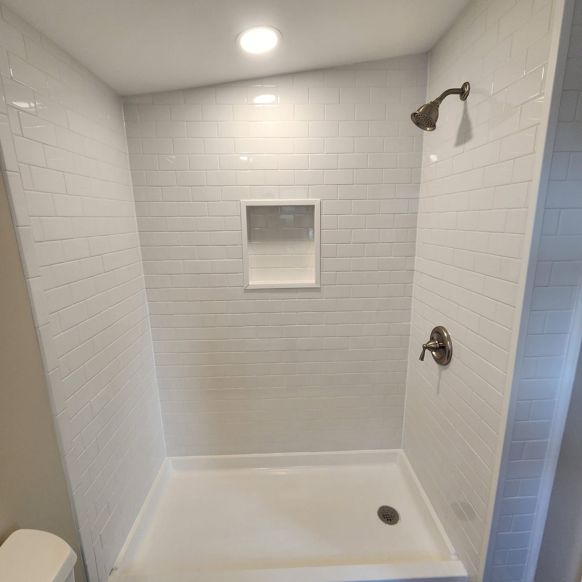 A shower stall with white tiles and a toilet in a bathroom.