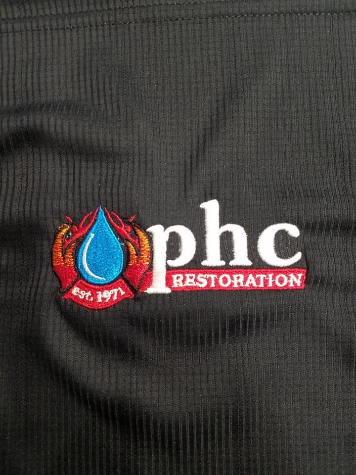The logo for phc restoration is embroidered on a black shirt.