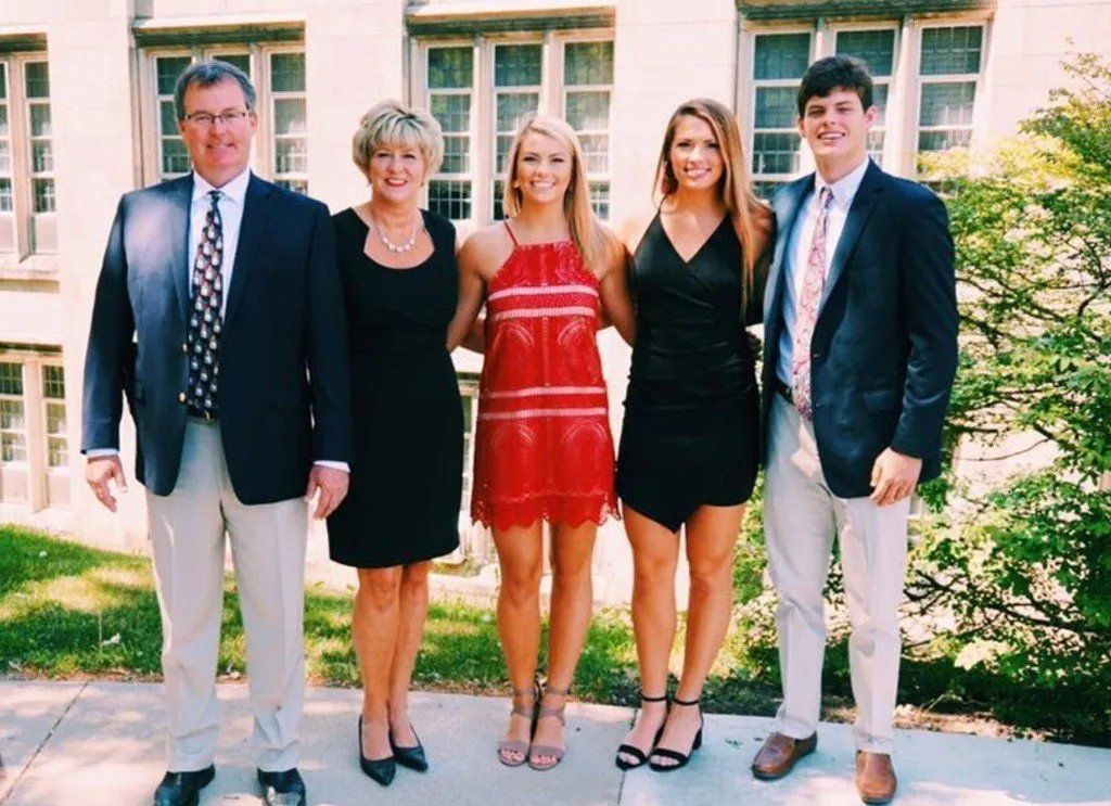 Family of five poses outside, smiling. Two women in dresses, two men in blazers, building in background.