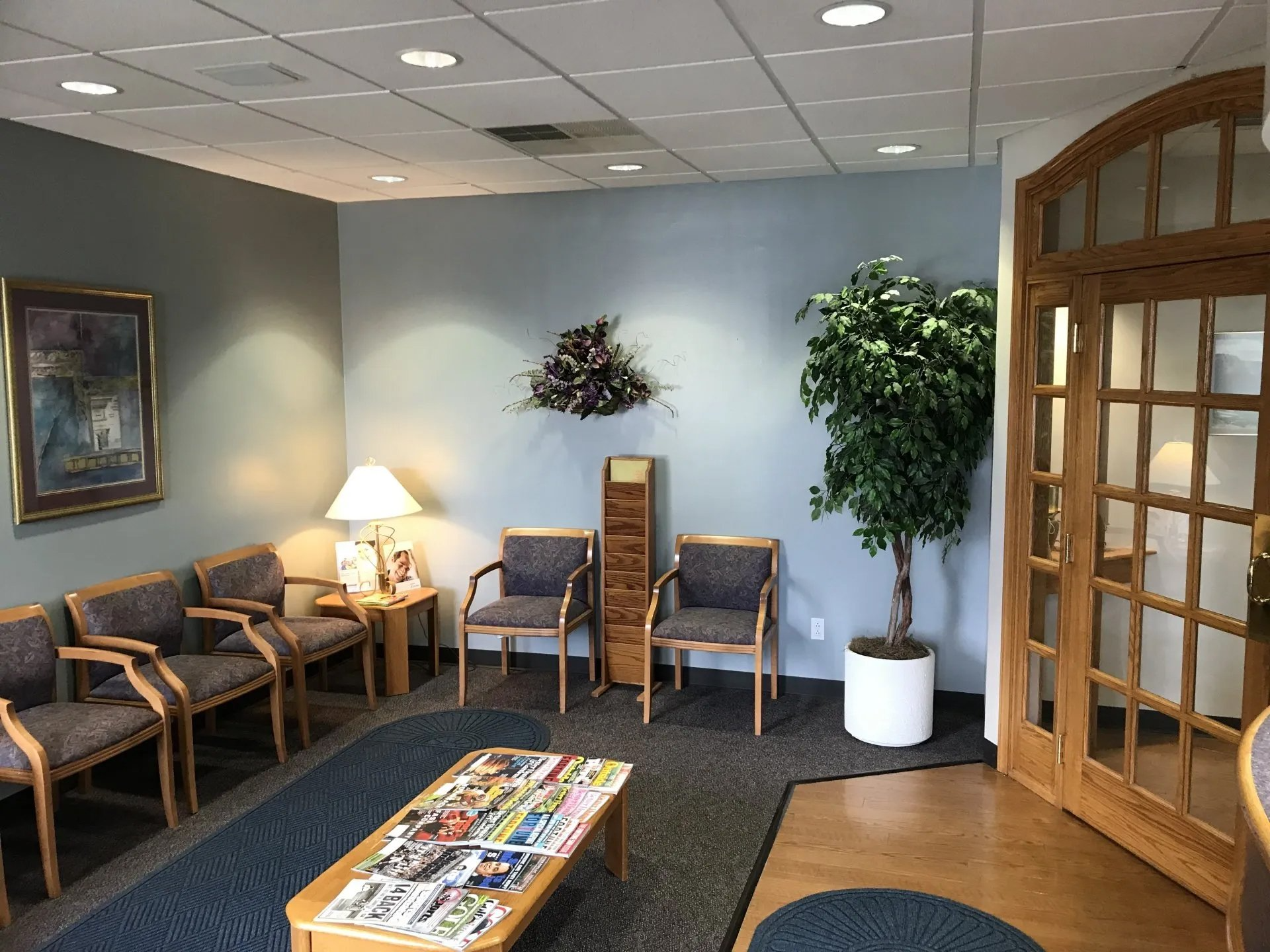 Waiting room with blue walls, chairs, and magazines. A potted tree sits near a wooden door.