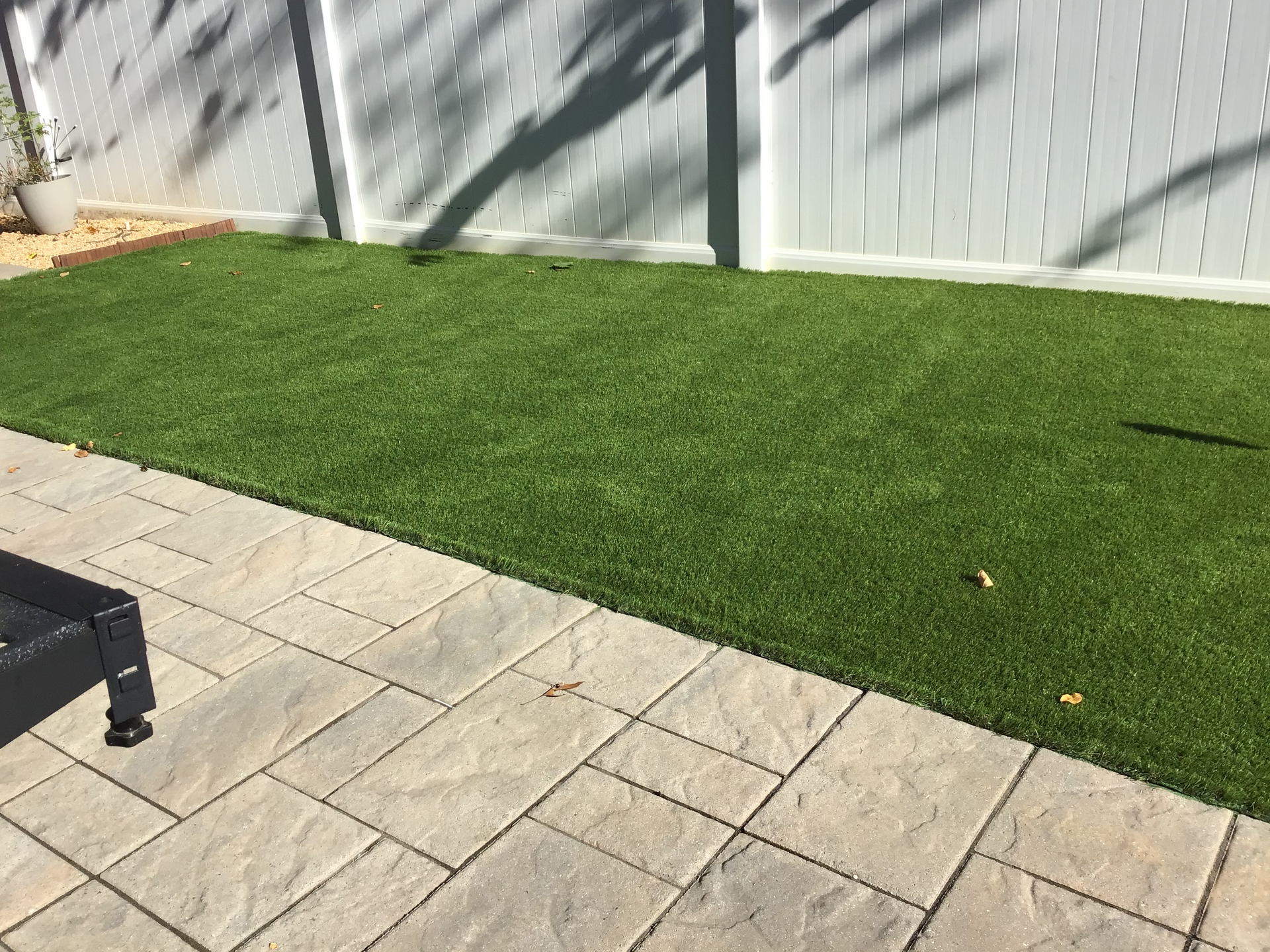 Patio with tan pavers next to green artificial turf, white fence in the background.