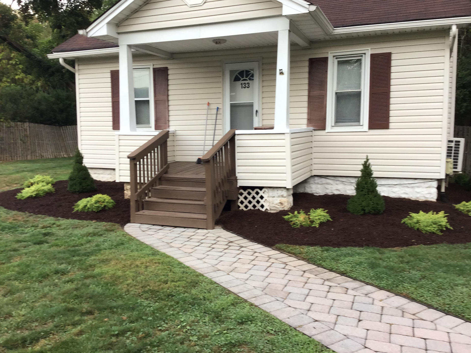 Small beige house with brown shutters, porch, and a brick pathway leading to the front door.
