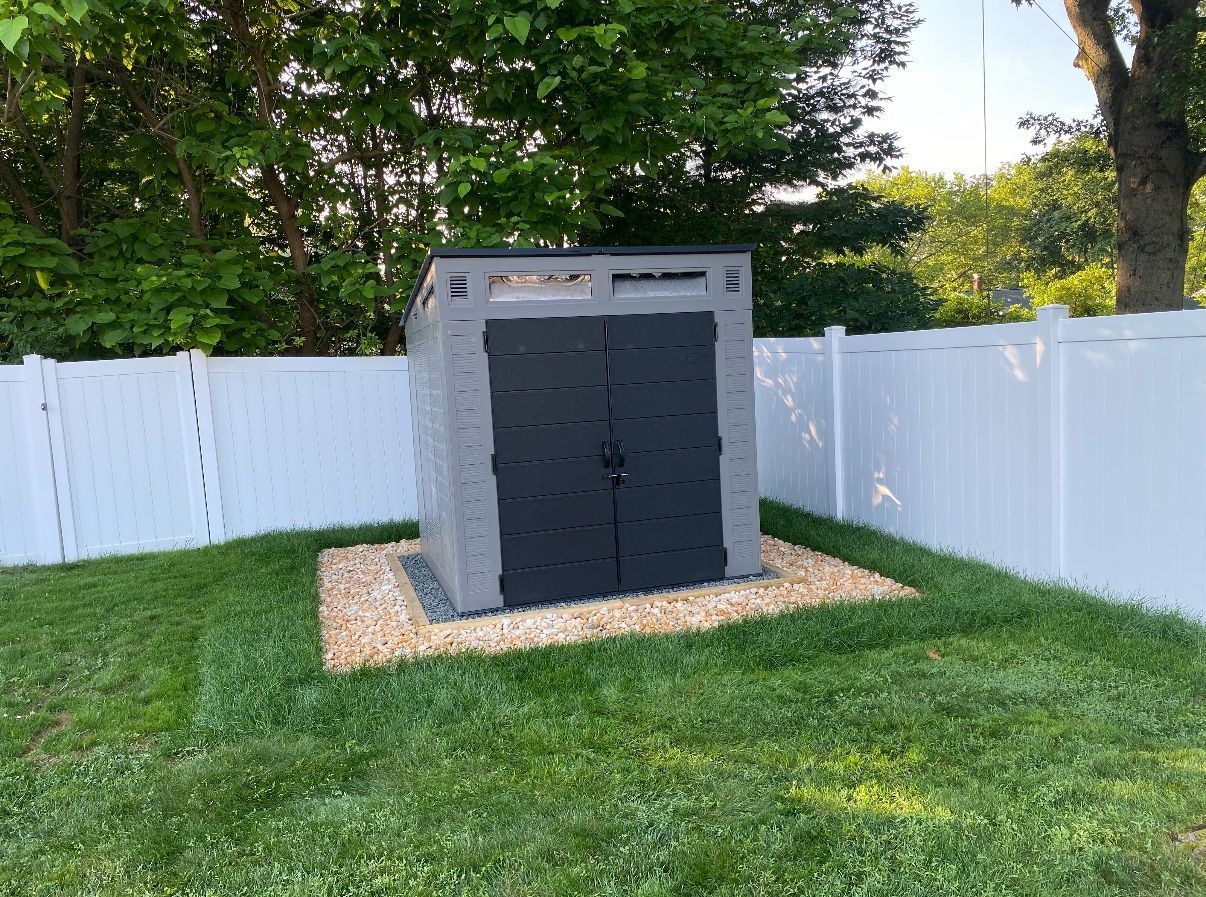 Storage shed with gray walls and black doors on a gravel base, surrounded by a white fence and grass.