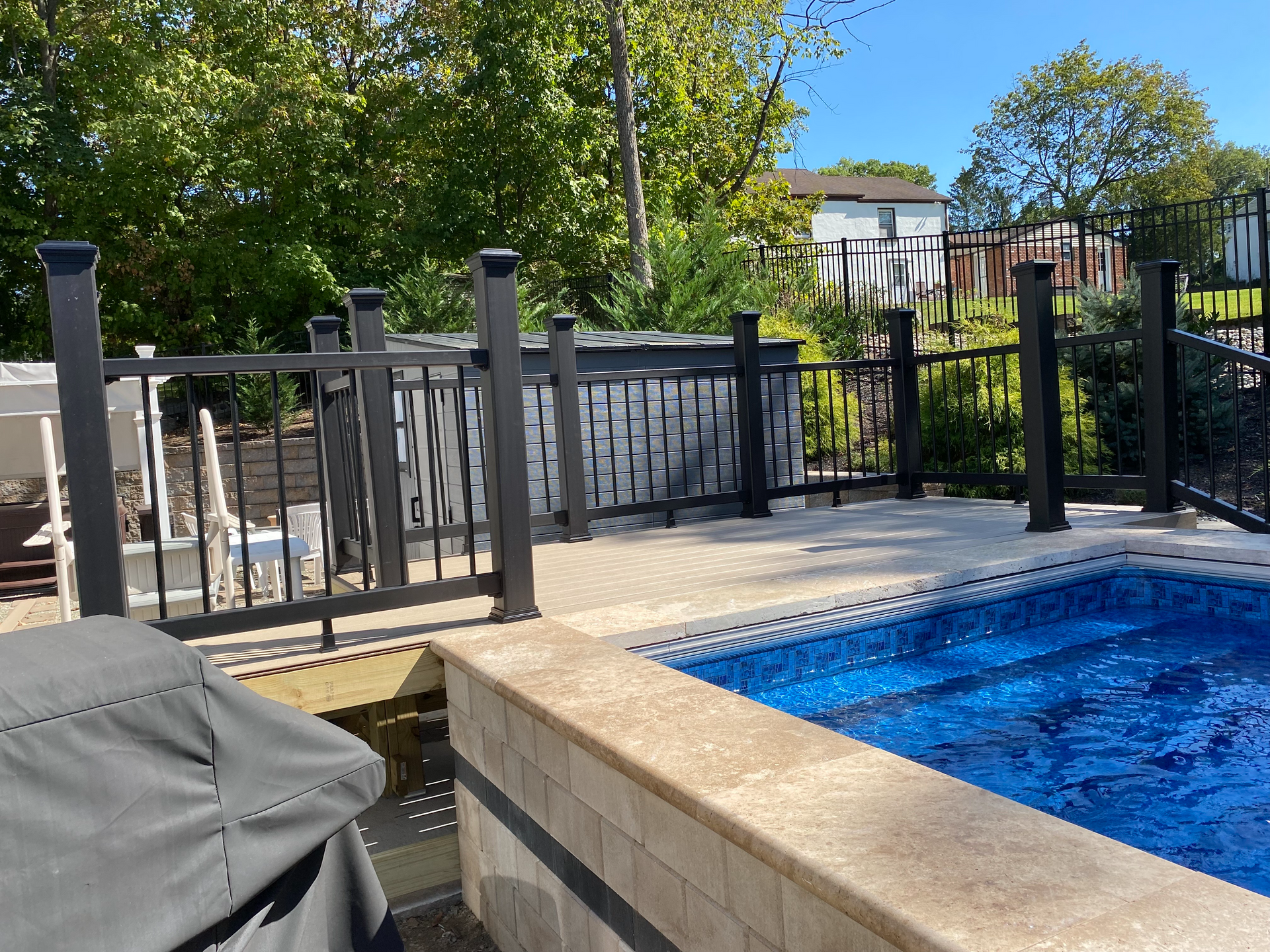 Backyard pool with black fence and a hot tub. Blue water, stone patio, sunny day.