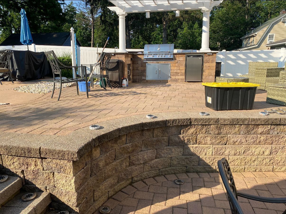 Brick patio with outdoor kitchen, pergola, and black and yellow storage bin.