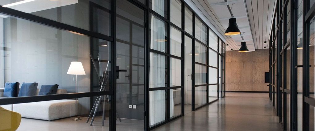 Modern office hallway with glass partitions, black frames, pendant lights, and a lounge area with a white sofa