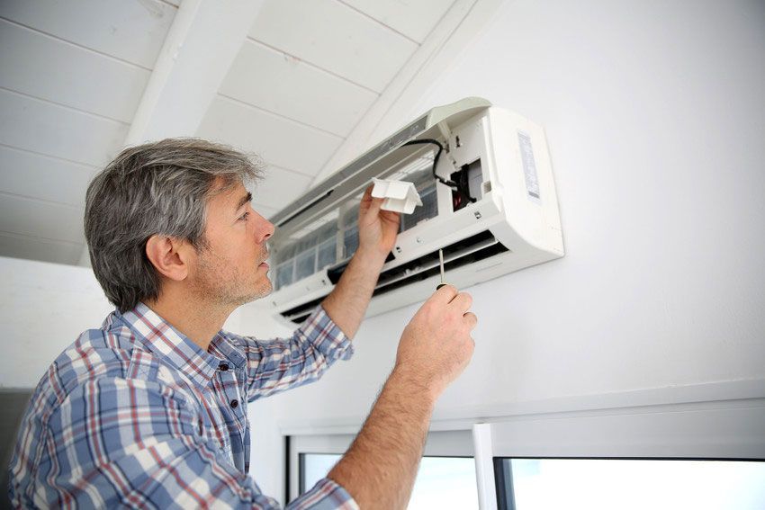 Man in plaid shirt repairs wall-mounted air conditioner with a screwdriver. White room setting.
