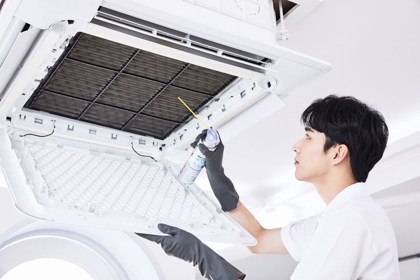 Person cleaning an air conditioning unit. Wearing gloves, spraying cleaner onto the filter, indoors.