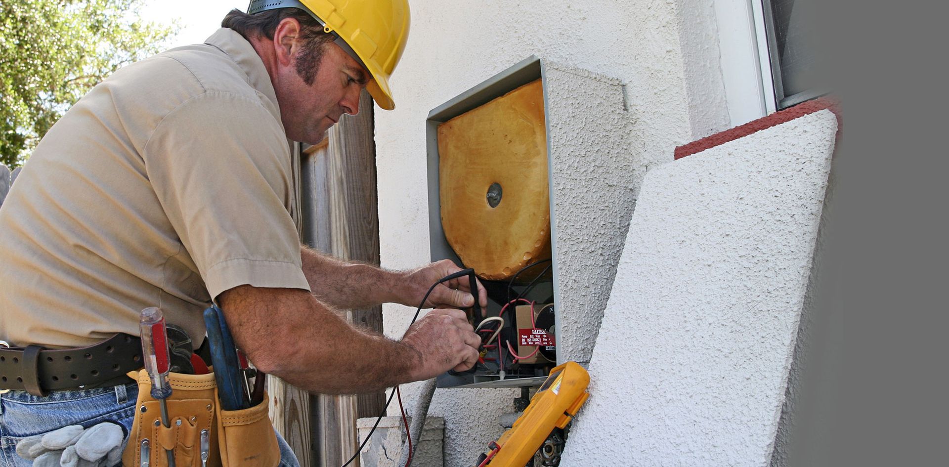 Electrician in a yellow hard hat tests electrical components in a gray box, with tools nearby.