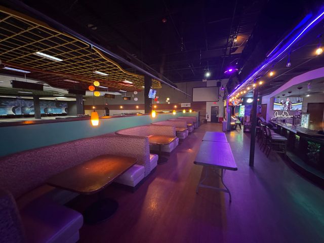 Empty modern bar/lounge with purple neon lighting, booth seating, and tables לאורך a long interior walkway