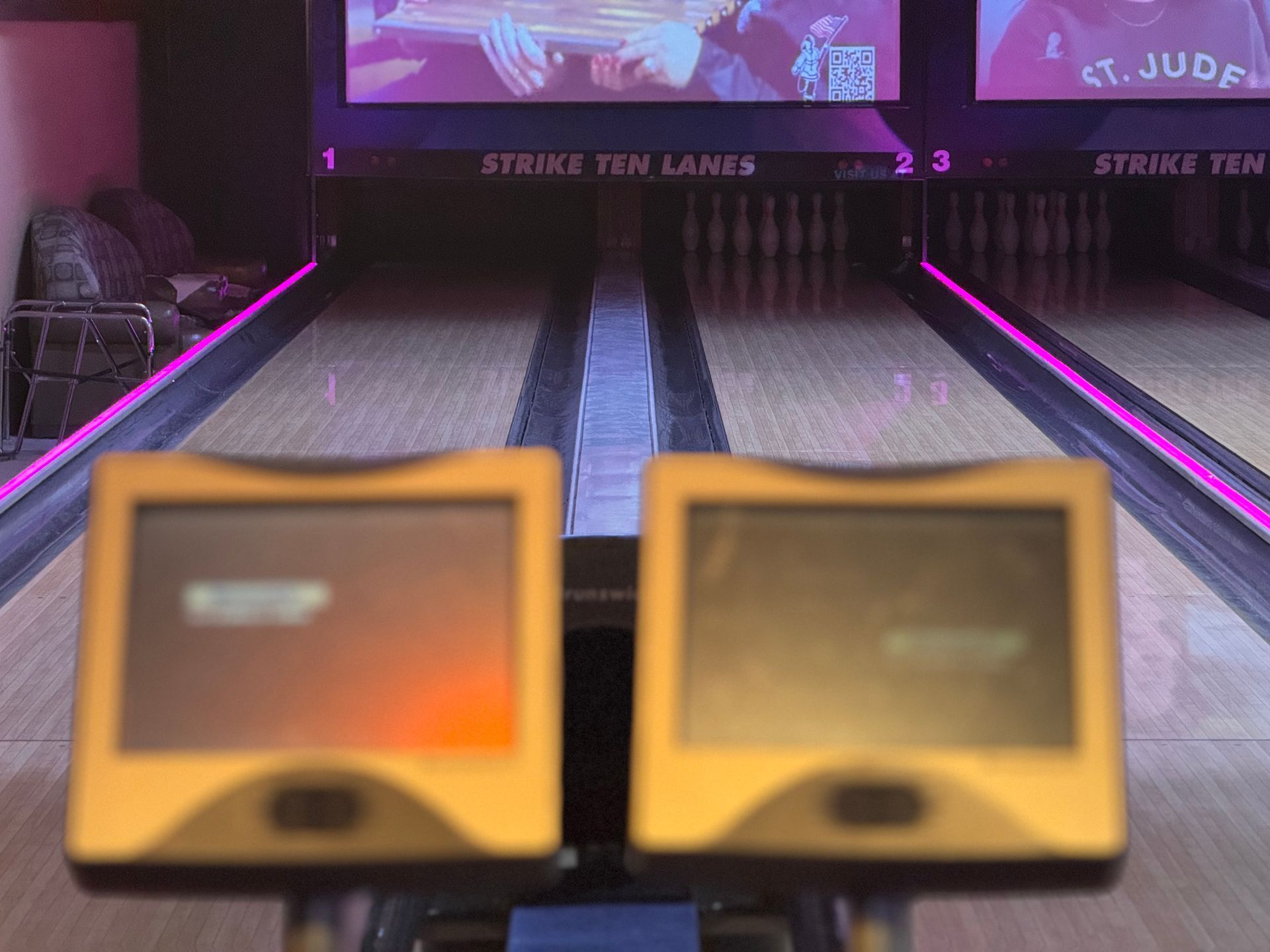 Bowling alley lanes lit in purple, viewed from behind two blurred scoring monitors