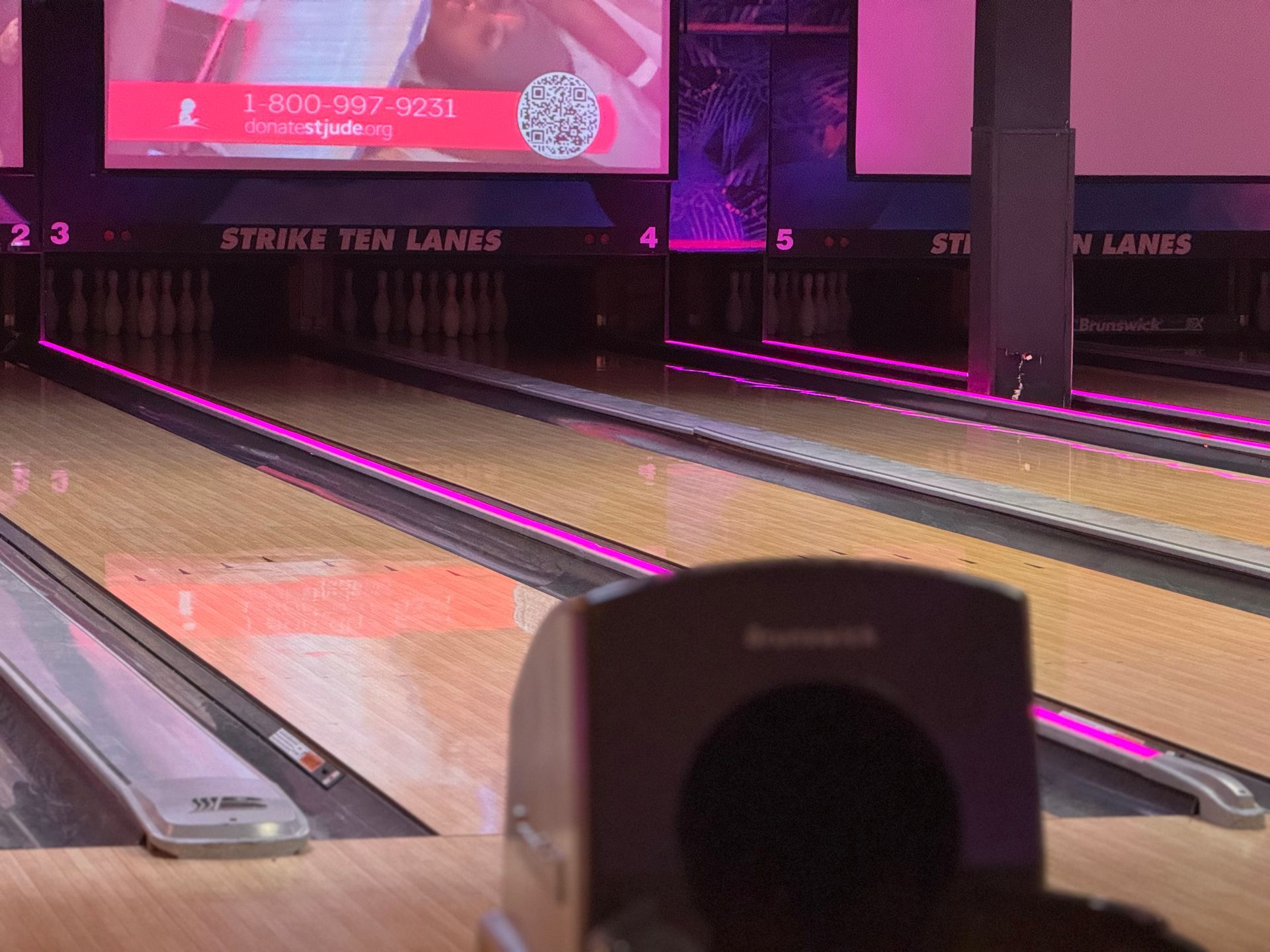Bowling alley lanes with bright pink lighting and a ball return in the foreground
