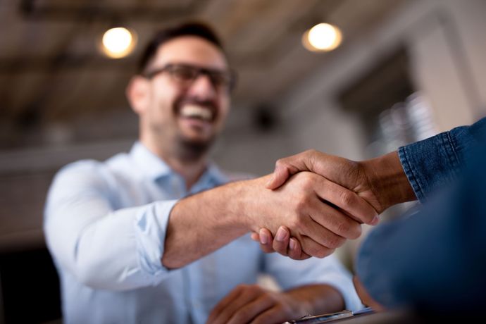Man in glasses smiles and shakes hands with another person. Interior shot, soft lighting.