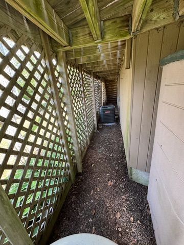 Narrow, dirt-floored walkway with latticework fencing, under a wooden deck. An AC unit is visible in the distance.