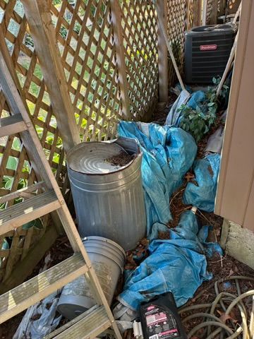 Cluttered outdoor space with a metal trash can, blue tarp, and air conditioner next to a latticework wall and ladder.