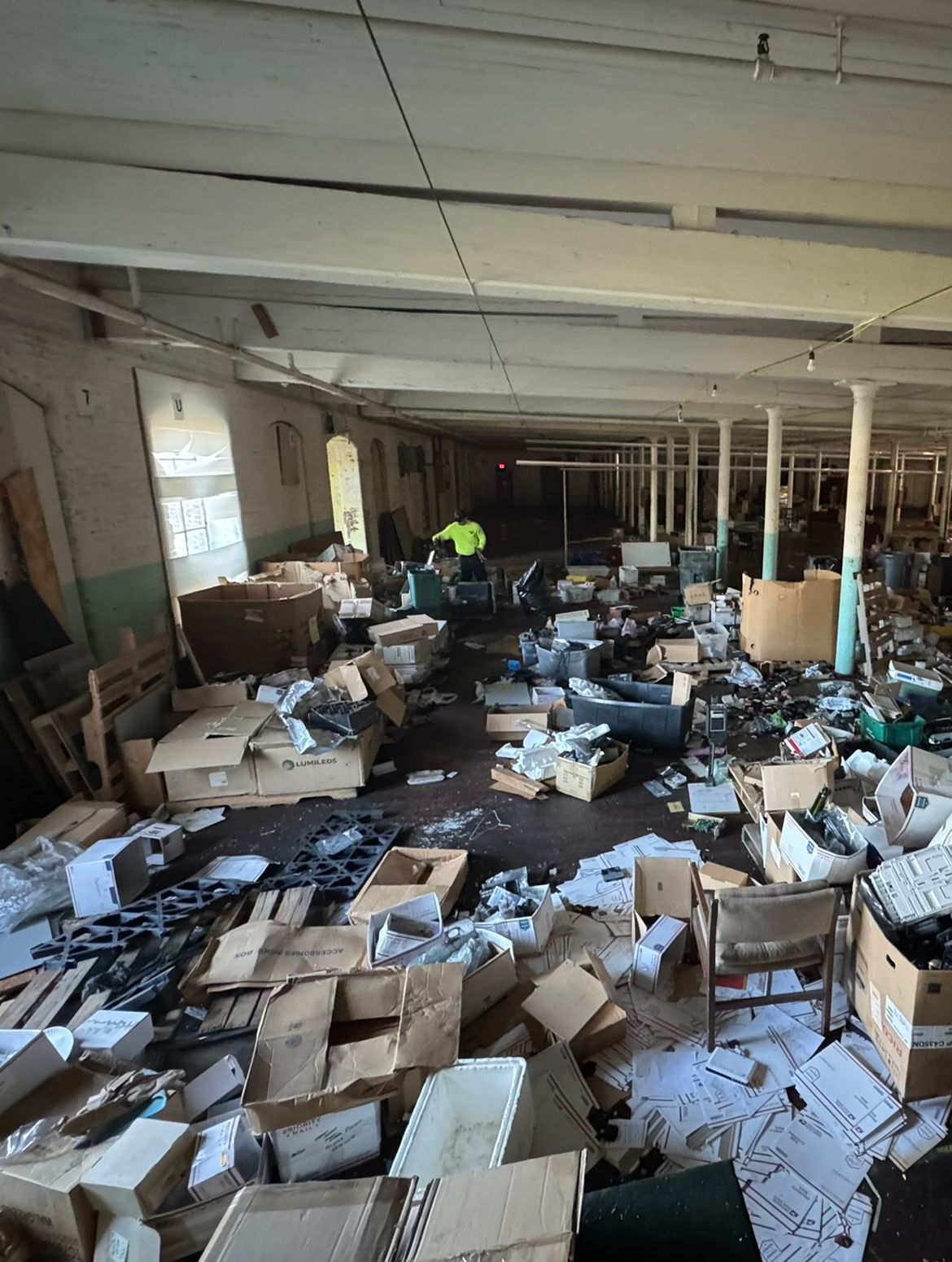 Messy interior with scattered boxes, debris, and a person in a yellow vest. Long, light-filled room.