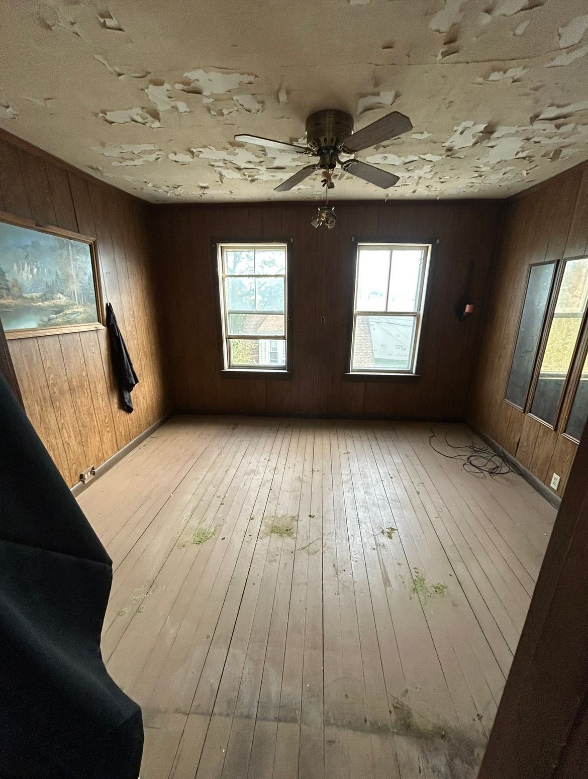 A room with peeling ceiling paint, wood panel walls, and two windows.